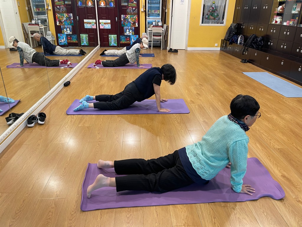 三人在紫色瑜伽墊上以手臂支撐身體並伸展腿部，左側牆面為鏡子，後方有置物櫃