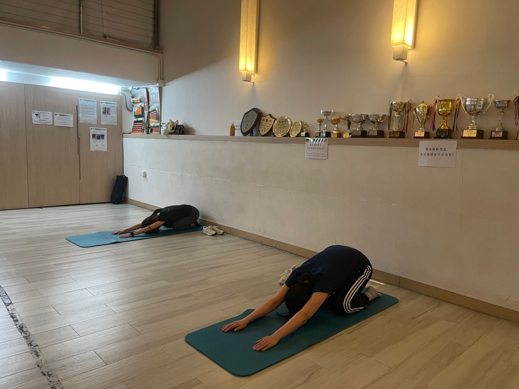 兩人在室內各自的瑜伽墊上採跪姿向前伸展，後方牆面展示多個獎盃與獎牌