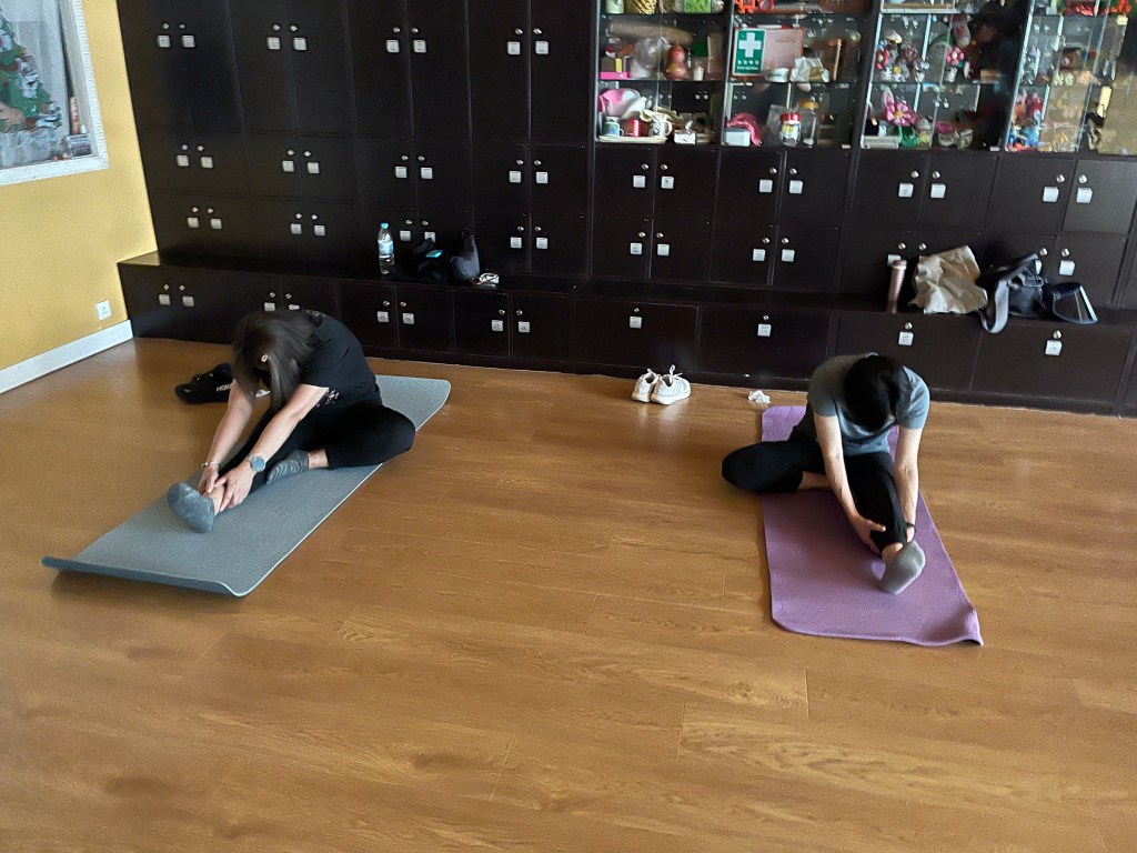 兩人分別坐在灰色和紫色瑜伽墊上，向前伸手觸碰伸直的腿部，背景有深色置物櫃