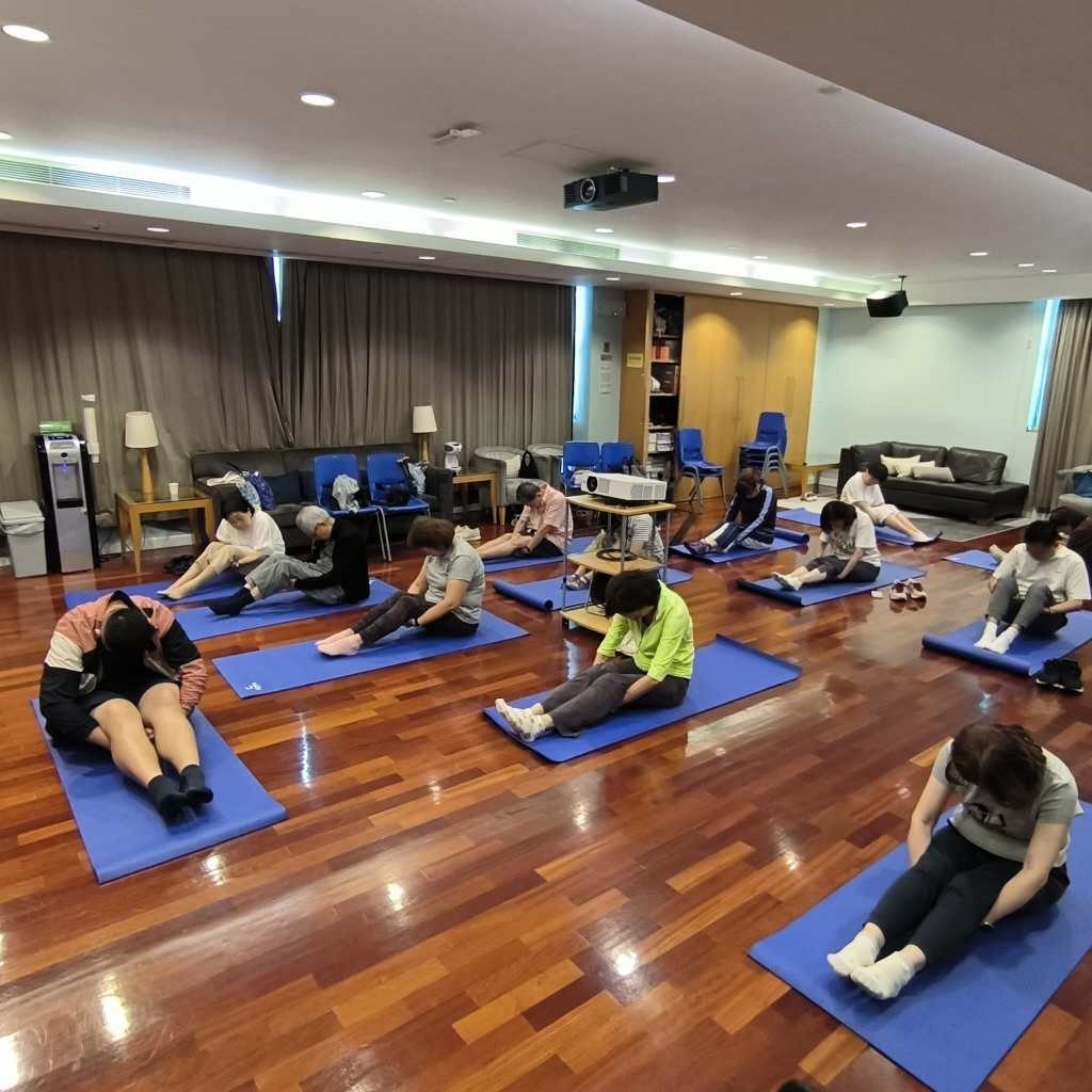 多人在室內藍色瑜伽墊上坐著伸展雙腿，周圍有椅子、沙發、投影機及窗簾